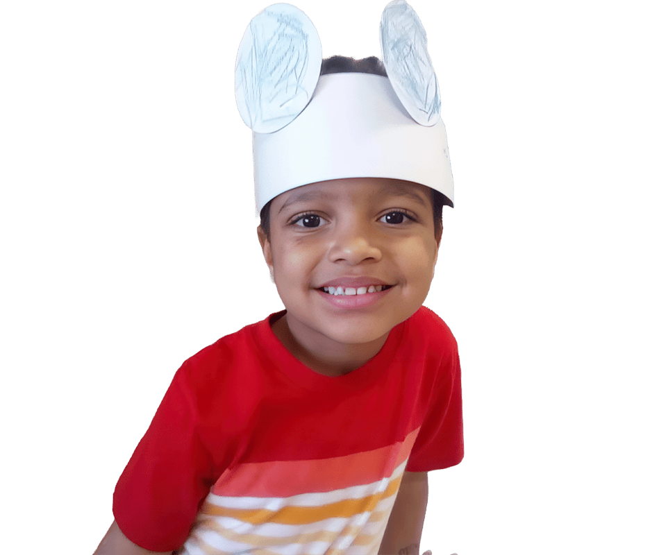 A young boy in a red shirt wearing a white paper crown with bunny ears smiling in front of a tree illustration.